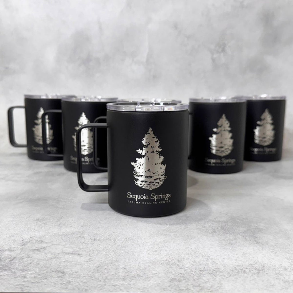 A black insulated mug with a clear lid and handle, featuring an engraved pine tree logo and the text 'Sequoia Springs' on the front, with five matching mugs arranged in the background on a light gray textured surface.