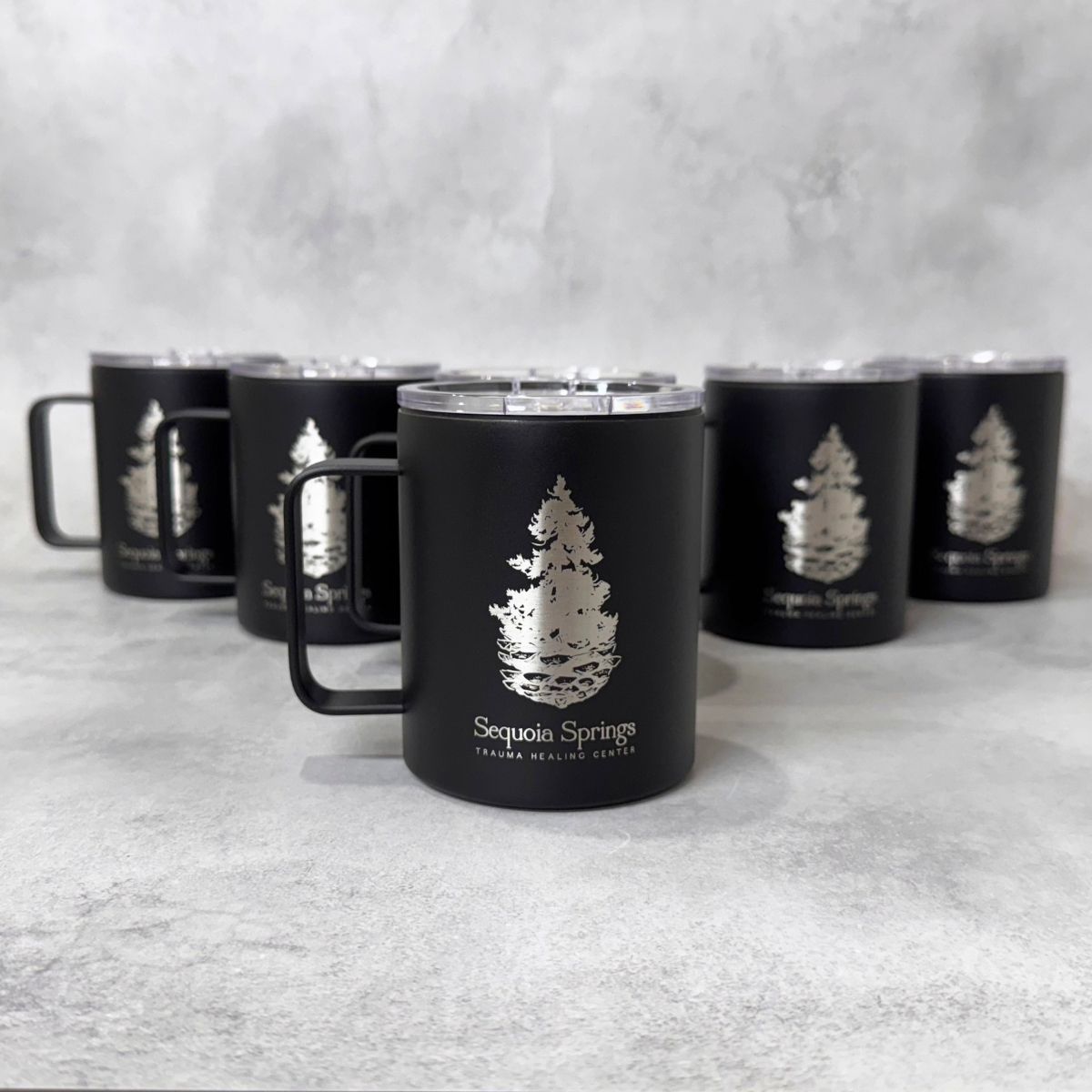 A black insulated mug with a clear lid and handle, featuring an engraved pine tree logo and the text 'Sequoia Springs' on the front, with five matching mugs arranged in the background on a light gray textured surface.