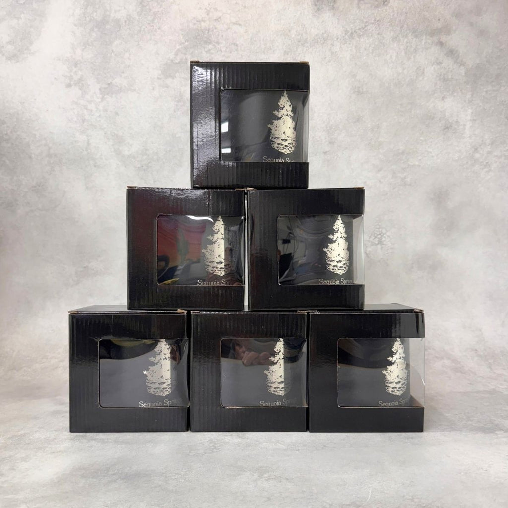Five black gift boxes stacked in a pyramid arrangement on a textured gray background. Each box has a clear window showing an engraved coffee mug inside.