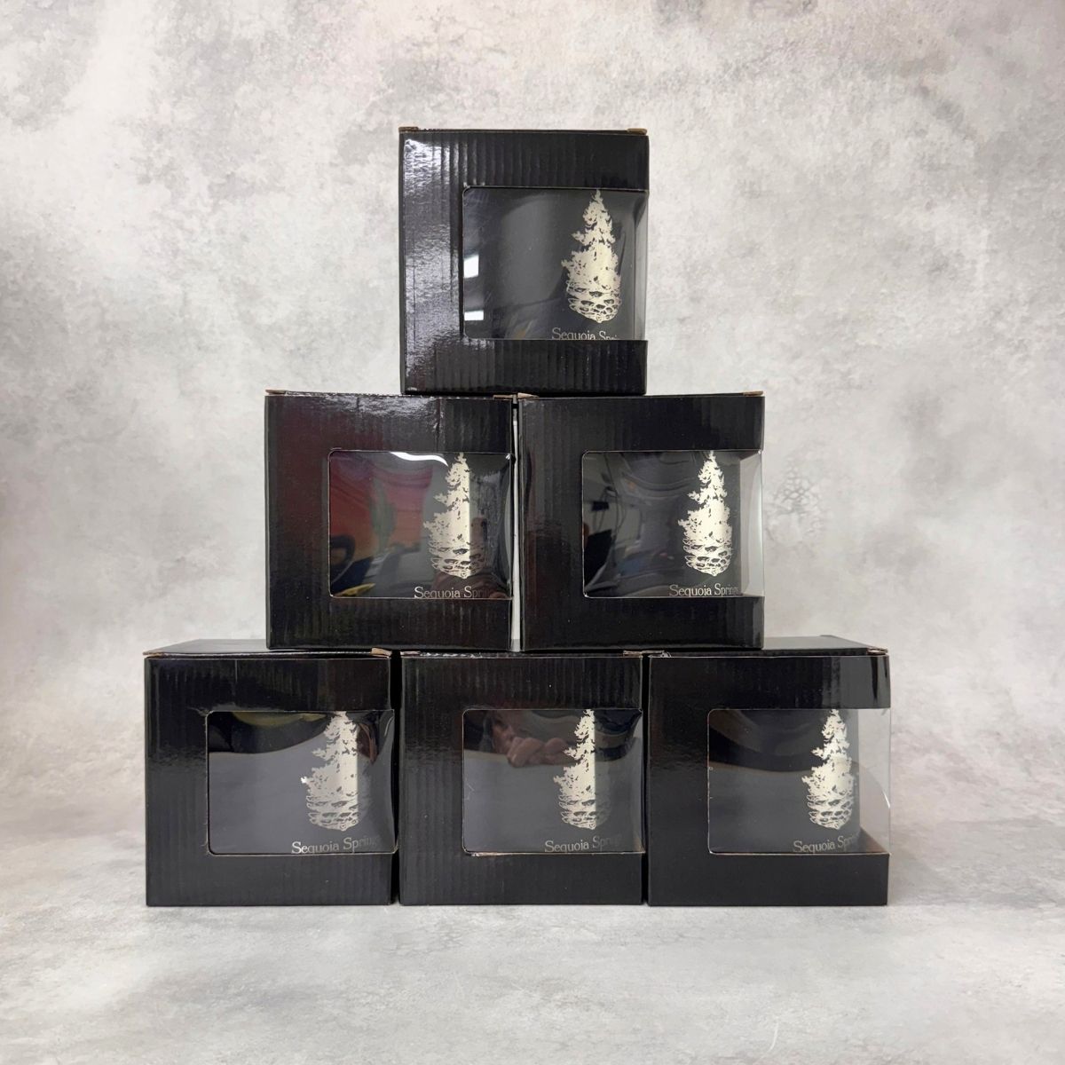 Five black gift boxes stacked in a pyramid arrangement on a textured gray background. Each box has a clear window showing an engraved coffee mug inside.
