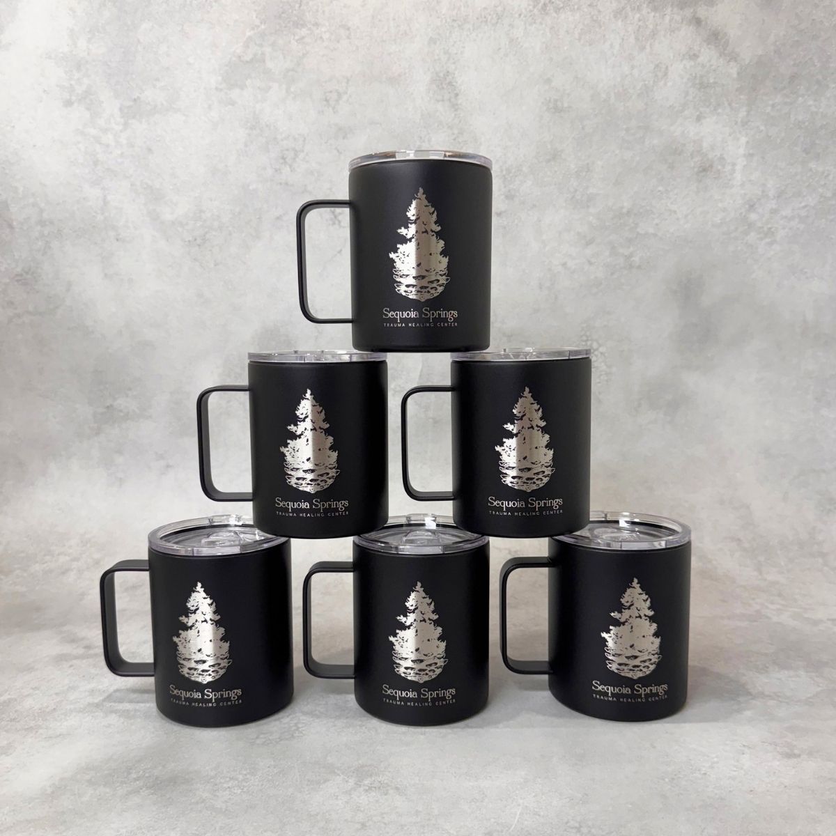 Six matte black insulated tumblers with clear lids are arranged in a pyramid on a light gray textured surface. Each tumbler has an engraved tree logo and the text 'Sequoia Springs.'