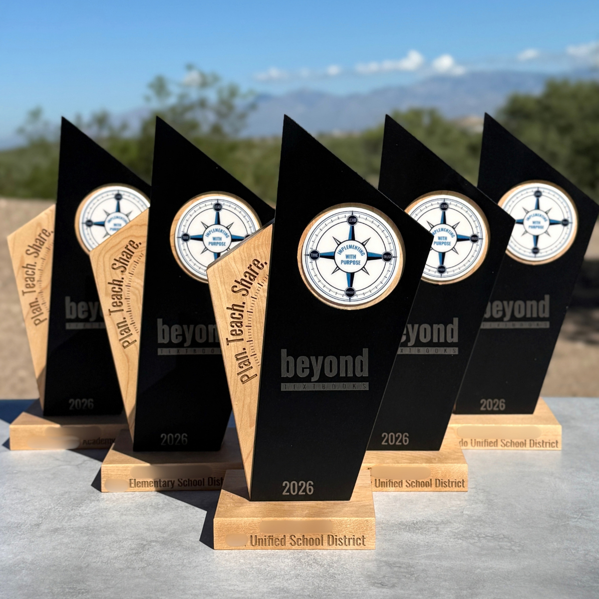 Six black and wood school district award plaques standing on a gray surface outdoors, with a blurred desert landscape and blue sky in the background. The plaques feature a circular emblem with a compass-like design and the words 'beyond' and '2026'.