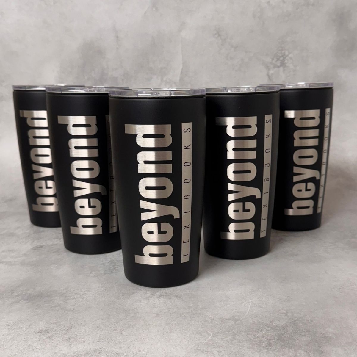 Five matte black insulated tumblers with clear lids arranged in a pyramid on a light gray textured surface. Each tumbler is printed vertically with the words "beyond" in large silver letters, with smaller text reading "TEXTBOOKS" running alongside.