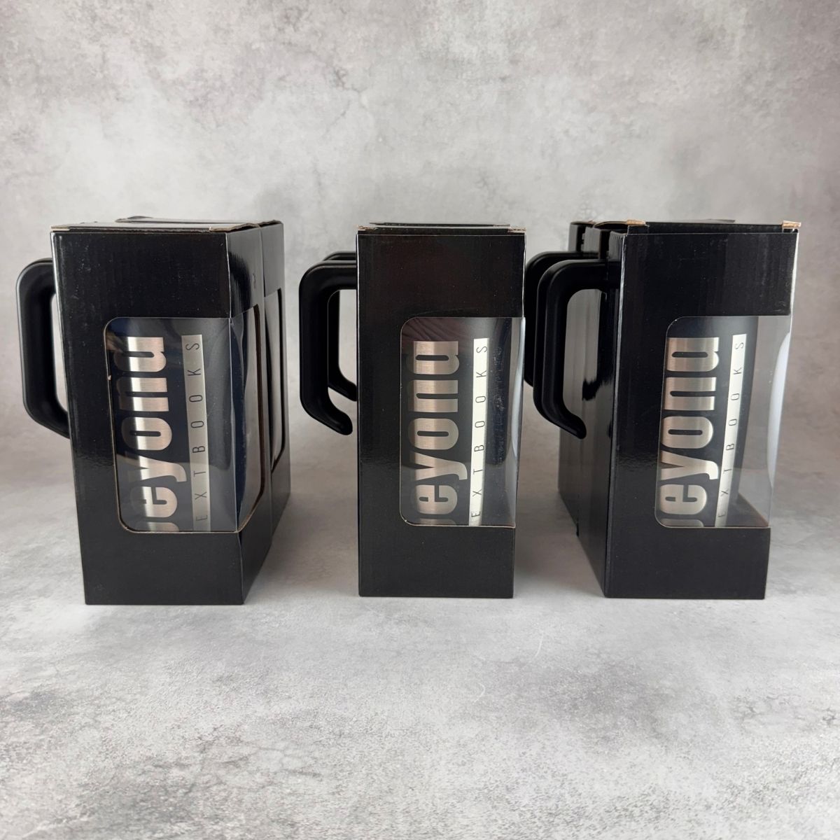 Three engraved black tumblers with handles, shown in their original black packaging boxes against a gray textured background.
