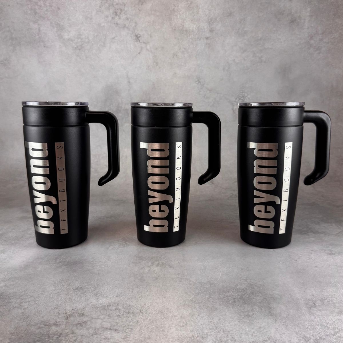 Three black insulated travel mugs with handles and lids, each engraved vertically with the word 'beyond' and smaller text reading 'textbooks', arranged side by side on a gray textured surface.