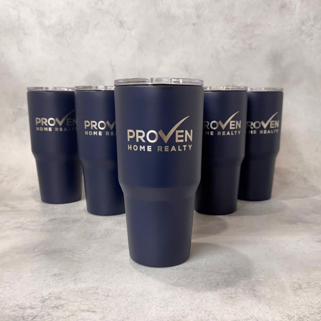 Five navy blue insulated tumblers with clear lids are arranged on a light gray textured surface. The front tumbler is centered and features the white text 'PROVEN HOME REALTY' with a checkmark logo, while the other tumblers show the same branding from different angles.