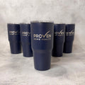 Five navy blue insulated tumblers with clear lids are arranged on a light gray textured surface. The front tumbler is centered and features the white text 'PROVEN HOME REALTY' with a checkmark logo, while the other tumblers show the same branding from different angles.