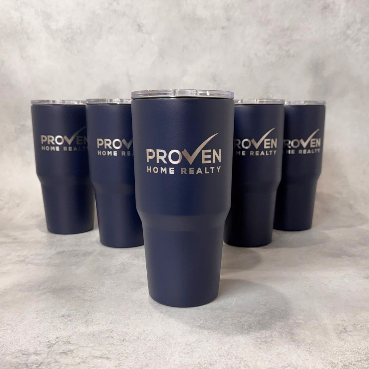 Five navy blue insulated tumblers with clear lids are arranged on a light gray textured surface. The front tumbler is centered and features the white text 'PROVEN HOME REALTY' with a checkmark logo, while the other tumblers show the same branding from different angles.