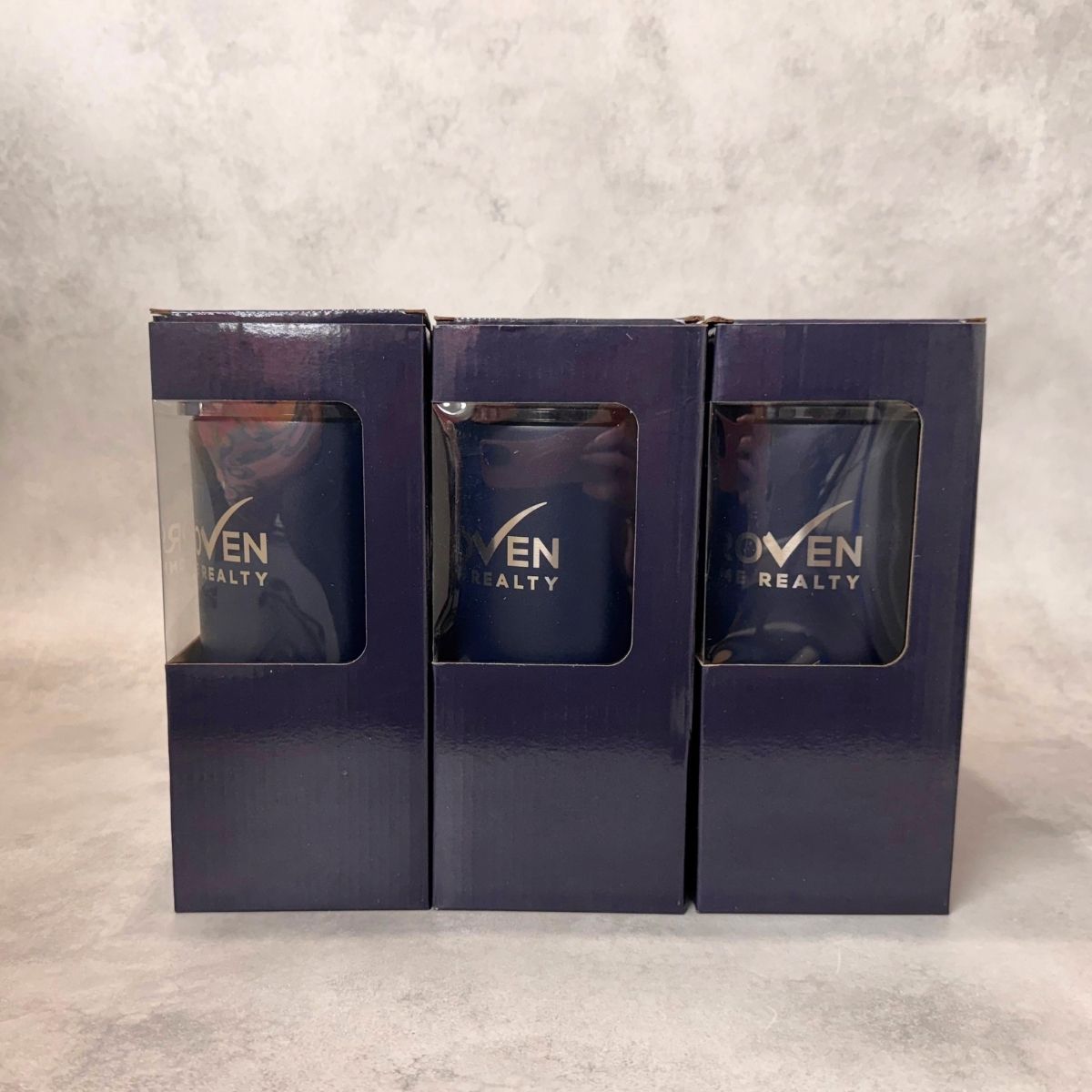 Three navy blue tumbler boxes with clear window cutouts, each displaying the 'Proven Home Realty' logo, arranged side-by-side in their matching dark navy retail packaging boxes against a textured gray background.