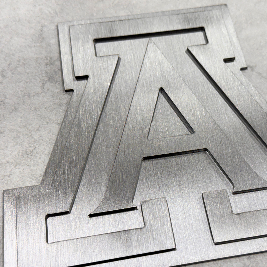 A close-up of a 2 layered brushed metal University of Arizona logo Block 'A' cutout with a brushed silver finish, resting on a textured light gray surface.