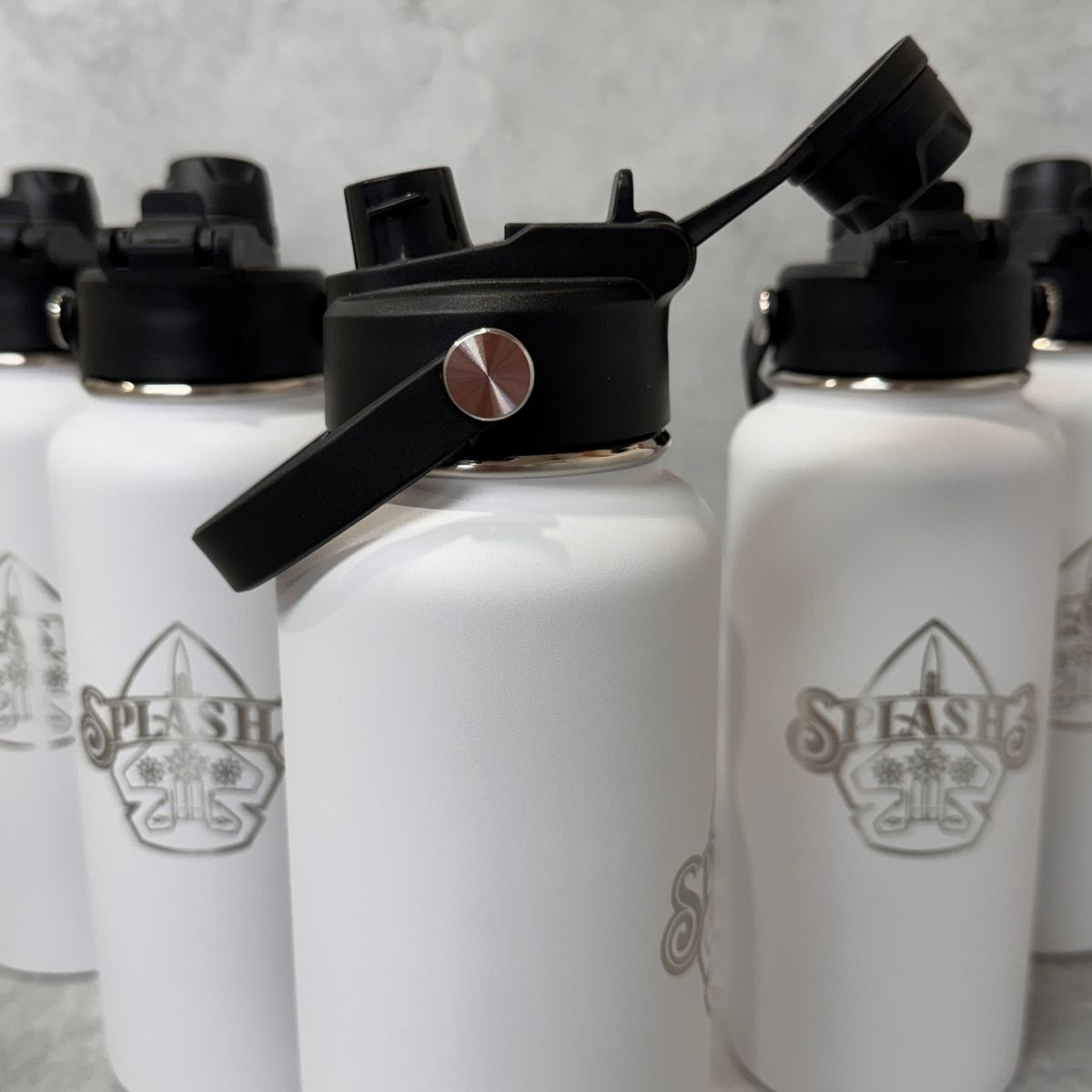 White insulated water bottles with black flip-straw lids and an engraved logo, arranged closely together on a light gray surface.