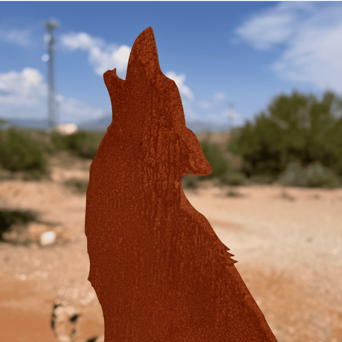 A rusted metal cutout of a wolf in profile, photographed outdoors against a blurred desert landscape with sand, shrubs, and blue sky.
