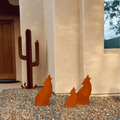 Three rust-colored metal howling coyote yard sculptures displayed on a gravel ground in front of a beige stucco house wall.