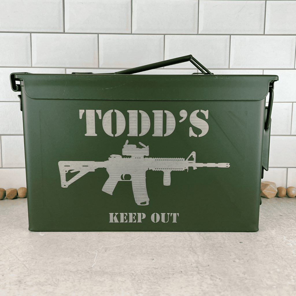 A green metal ammunition can with a handle, featuring an engraved stencil-style graphic of an AR-15 rifle and the text 'TODD'S KEEP OUT'. The can is set on a light gray concrete floor against a white subway tile wall.