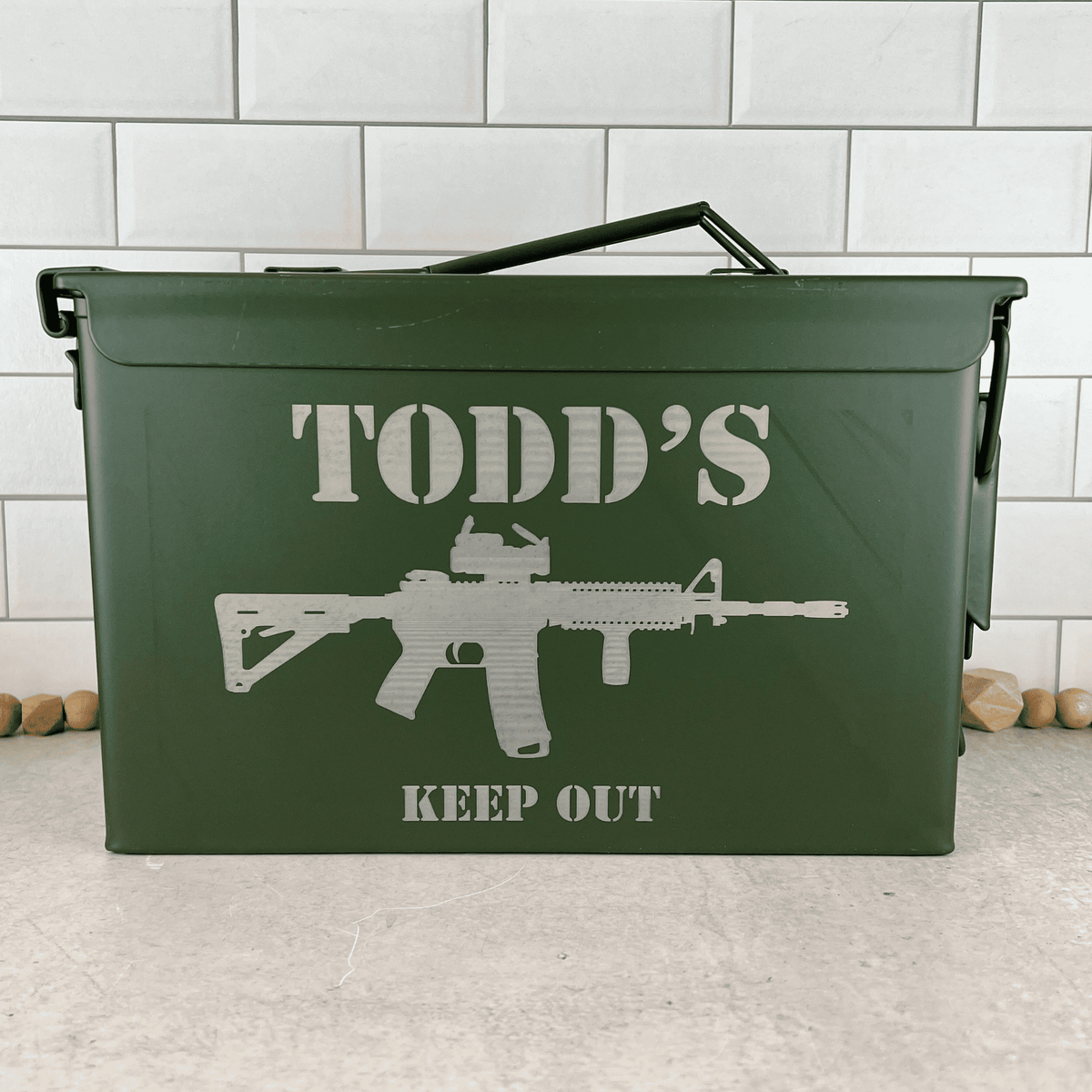 A green metal ammunition can with a handle, featuring an engraved stencil-style graphic of an AR-15 rifle and the text 'TODD'S KEEP OUT'. The can is set on a light gray concrete floor against a white subway tile wall.