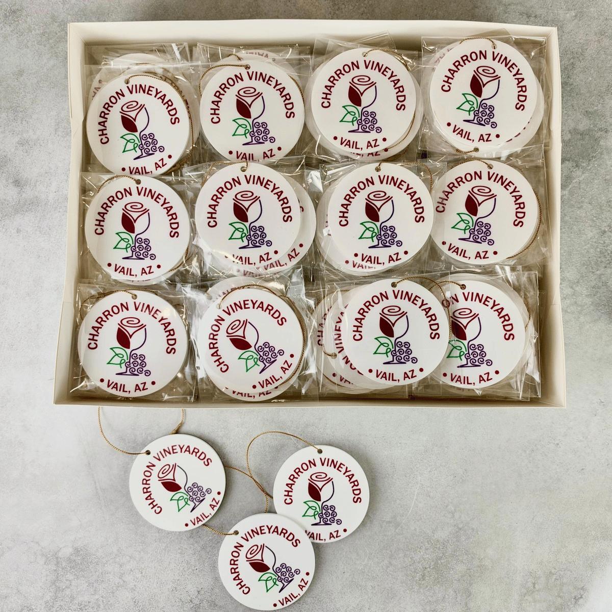 A boxed set of round white ornaments with printed 'Charron Vineyards' logos and grape illustrations, arranged in a tray with several additional ornaments placed in front on a light stone surface.
