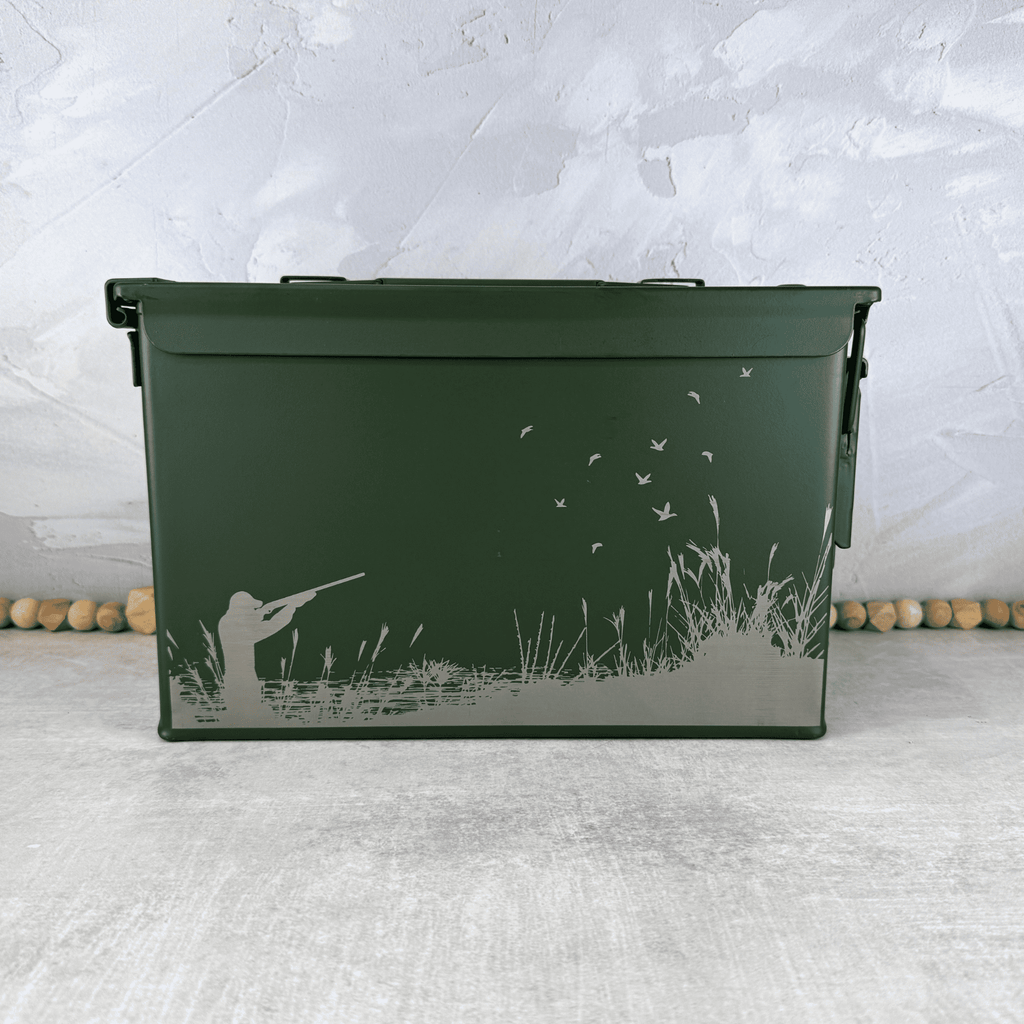 A dark green metal ammo storage box with a lid, decorated with an engraved graphic of a duck hunter silhouette, tall grass, and flying ducks, set against a textured gray background.