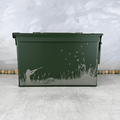 A dark green metal ammo storage box with a lid, decorated with an engraved graphic of a duck hunter silhouette, tall grass, and flying ducks, set against a textured gray background.
