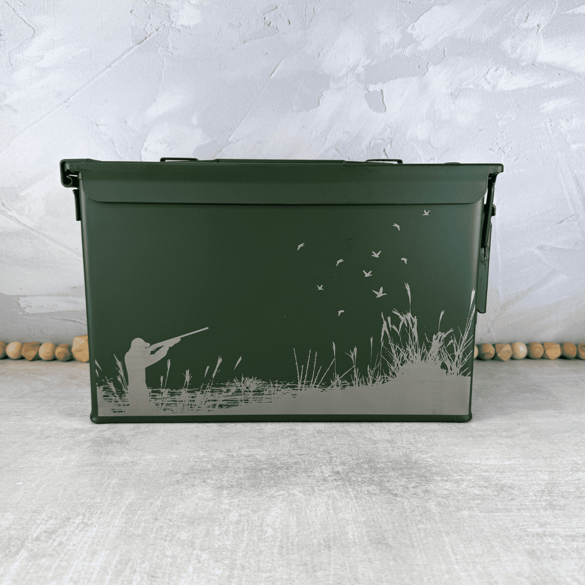 A dark green metal ammo storage box with a lid, decorated with an engraved graphic of a duck hunter silhouette, tall grass, and flying ducks, set against a textured gray background.