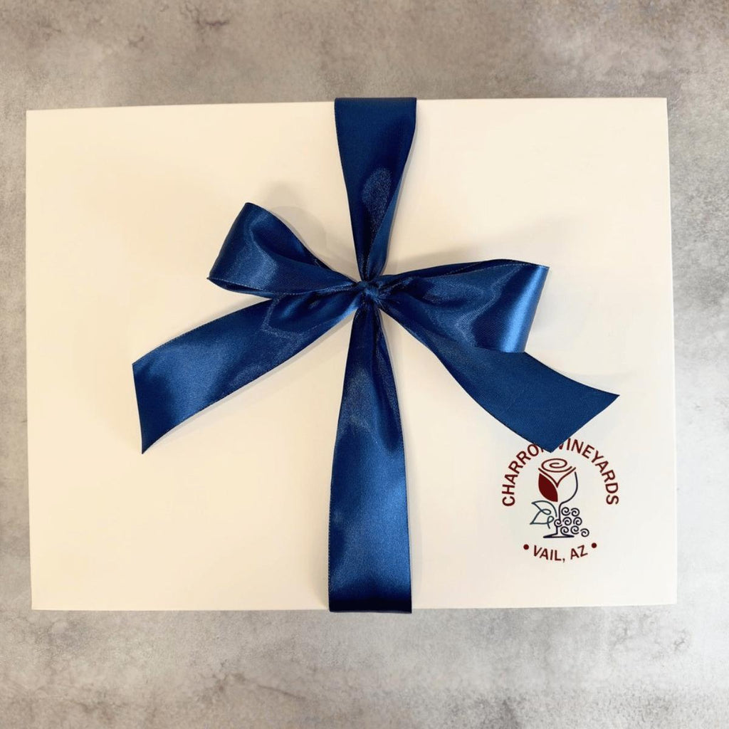 A square white custom printed gift box tied with a large dark blue satin ribbon bow, placed on a light gray textured surface. The box has a circular red and black logo for 'Charro's Vineyards' in the bottom right corner.