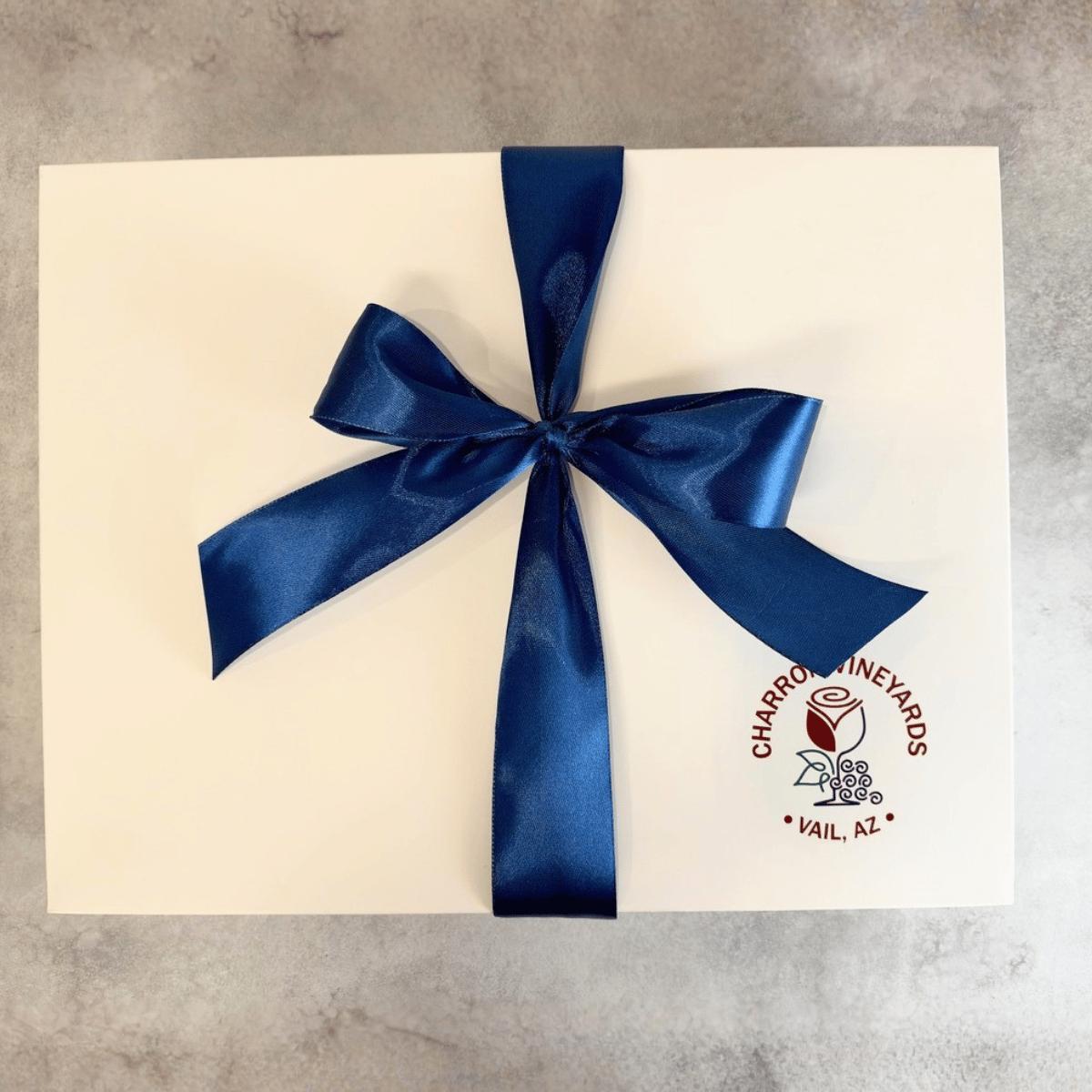 A square white custom printed gift box tied with a large dark blue satin ribbon bow, placed on a light gray textured surface. The box has a circular red and black logo for 'Charro's Vineyards' in the bottom right corner.