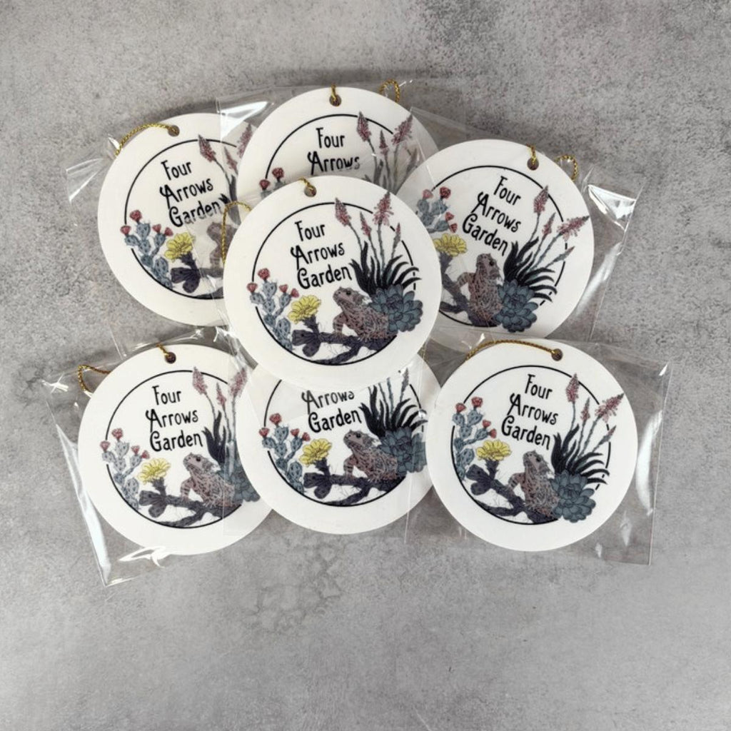 Seven round white hanging custom business ornaments in clear plastic packaging, printed with the phrase 'Four Arrows Garden' and illustrated with birds, flowers, and foliage, arranged on a gray textured surface.