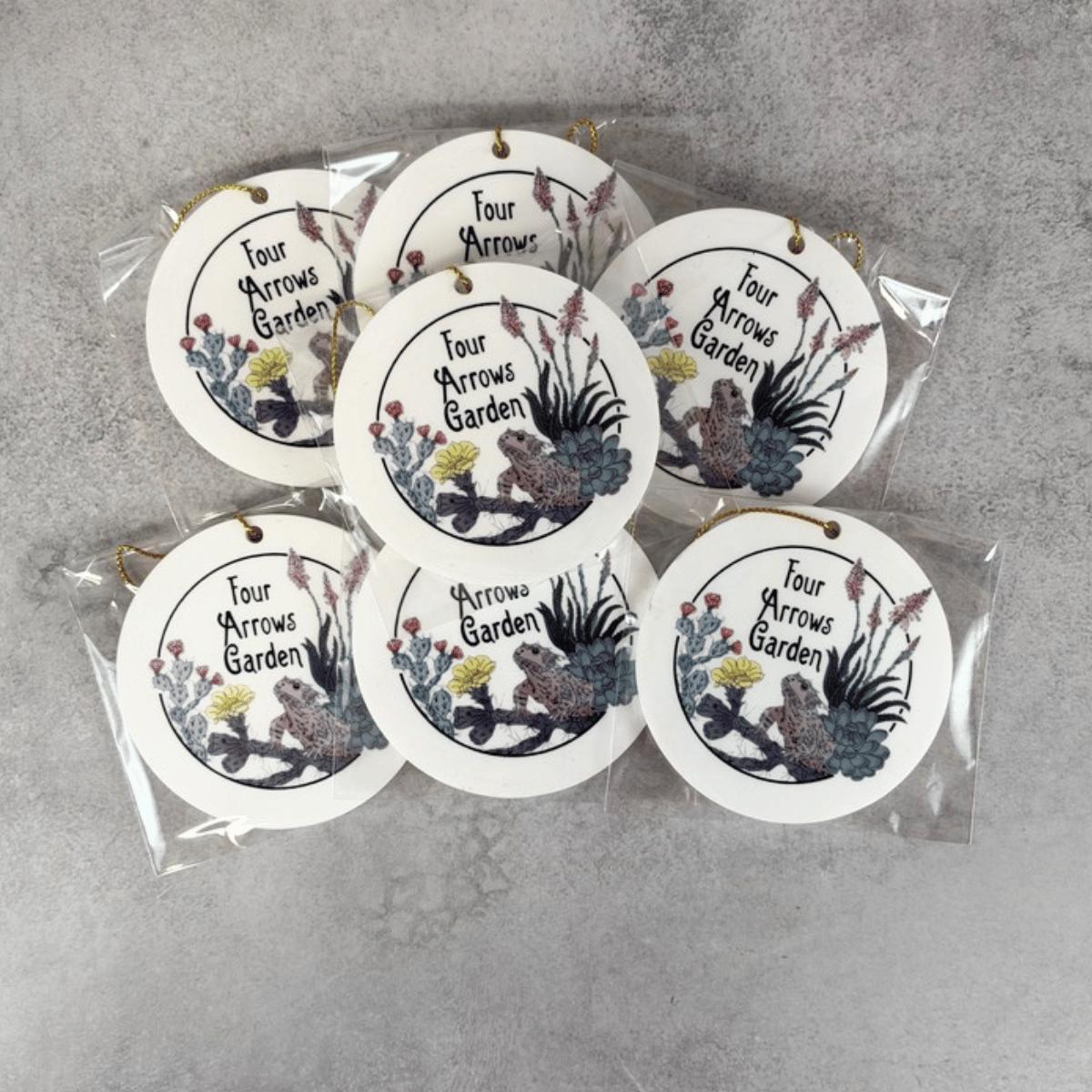 Seven round white hanging custom business ornaments in clear plastic packaging, printed with the phrase 'Four Arrows Garden' and illustrated with birds, flowers, and foliage, arranged on a gray textured surface.