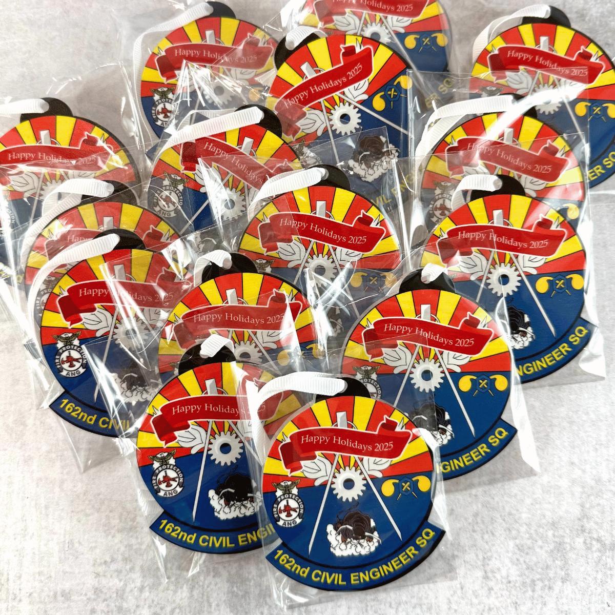 Multiple small packaged custom holiday ornaments arranged together on a light surface. Each item is wrapped in clear plastic and shows a red, yellow, blue, white, and black design with the text 'Happy Holidays 2025' and '162nd Civil Engineer SQ'.