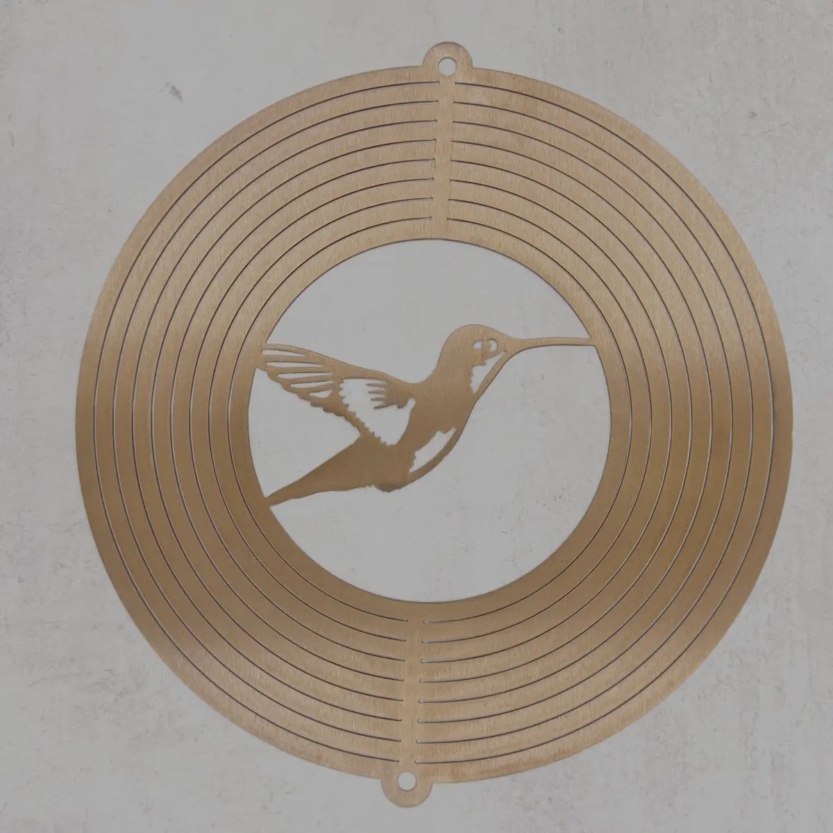 A circular metal wall art piece featuring concentric rings and a central hummingbird silhouette, shown against a textured light gray background.