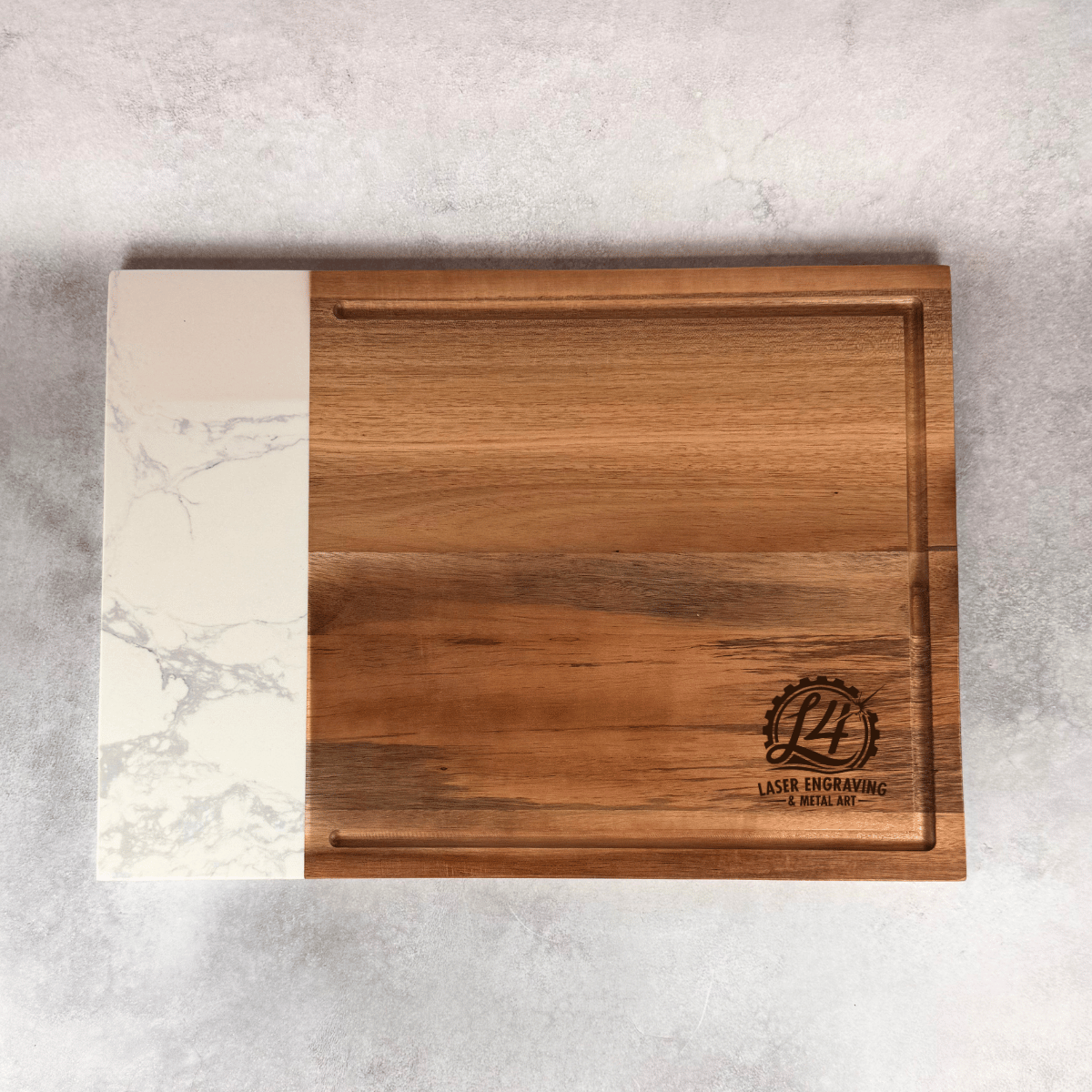 A rectangular wooden cutting board with a white marble-patterned left edge, photographed from above on a light gray textured surface. A circular laser-engraved logo reading 'L4 LASER ENGRAVING' is visible near the lower right corner.