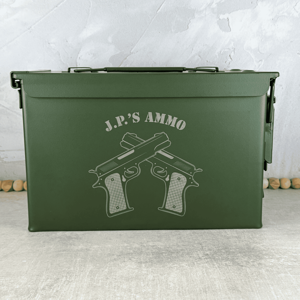 An olive green metal ammunition storage box with a handle, featuring an engraved printed graphic of two crossed handguns and the text 'J.P.'s Ammo.'