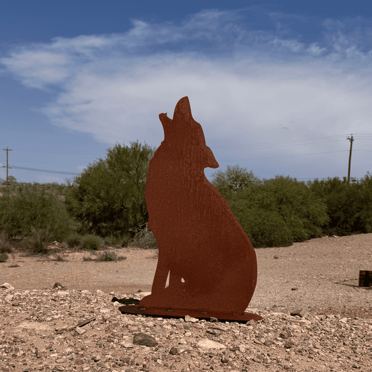 A rust-colored metal art piece of a howling coyote standing on a gravelly ground in an outdoor desert landscape with shrubs, trees, and a blue sky.