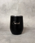 A matte black insulated stemless wine tumbler with a clear lid and the name 'Sarah' printed in elegant silver script on the front, set against a textured gray background.