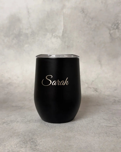 A matte black insulated stemless wine tumbler with a clear lid and the name 'Sarah' printed in elegant silver script on the front, set against a textured gray background.