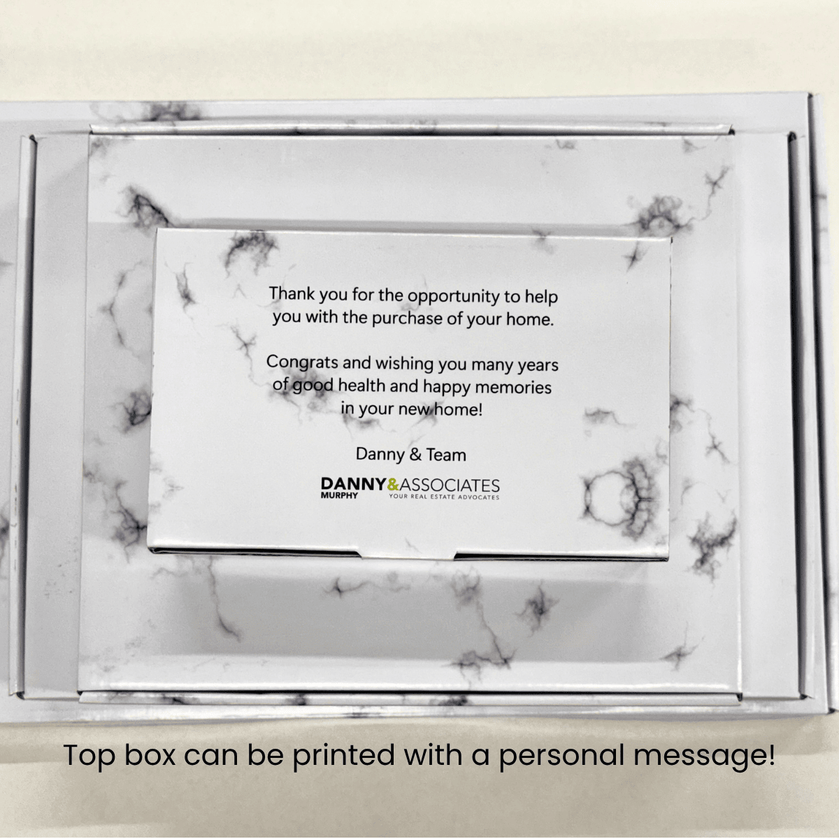 A white marble-patterned keepsake box with a printed message on the top. The card reads a congratulatory message for a home purchase and includes the brand name Danny & Associates.