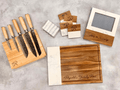 A coordinated kitchen gift arranged on a light gray textured surface, including a wood knife block with five knives, a large wood cutting board with a white marble vertical panel, a small photo frame with a wood base, and several matching coasters with monogram initials.