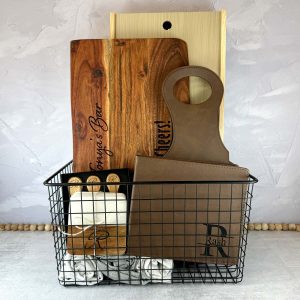 wire black metal basket with engraved cutting board, engraved BBQ set, and engraved coasters and charcuterie utensils inside. Basket also has rolled up dish towels in the bottom.