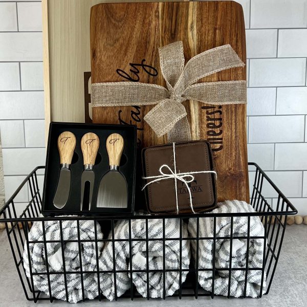 wire black metal basket with engraved cutting board, engraved BBQ set, and engraved coasters and charcuterie utensils inside. Basket also has rolled up dish towels in the bottom.