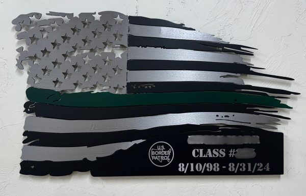 silver, black, and thin green line metal flag with room for engraving at the bottom. Flag is hung on a white brick wall.